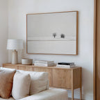 A minimalist living room features a light wood sideboard, white table lamp, stacked books, and a bowl. Above hangs the Winter Silence Canvas Art depicting a snowy landscape with two bare trees. Neutral tones complete the decor.