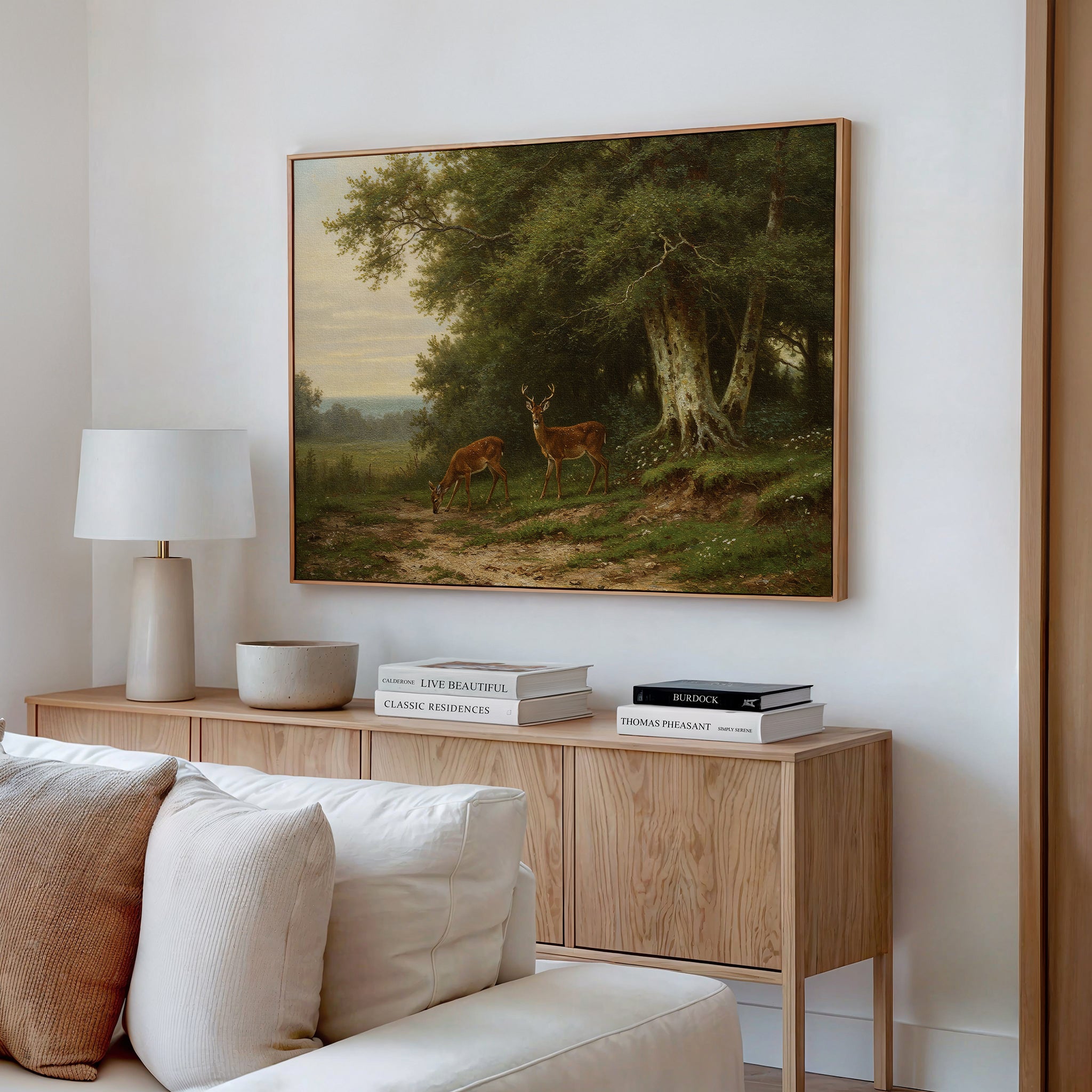 A cozy living room features a light wood sideboard, lamp, décor, books, and the Woodland Watch Canvas Art—a framed painting of two deer grazing by trees—perfect for nature lovers and beautifully displayed above the sideboard.