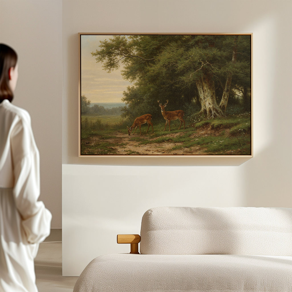 A woman in a white robe stands in a softly lit room, gazing at the Woodland Watch Canvas Art—a framed painting of two deer in a forest—hanging above a beige sofa or bed, ideal décor for nature lovers.