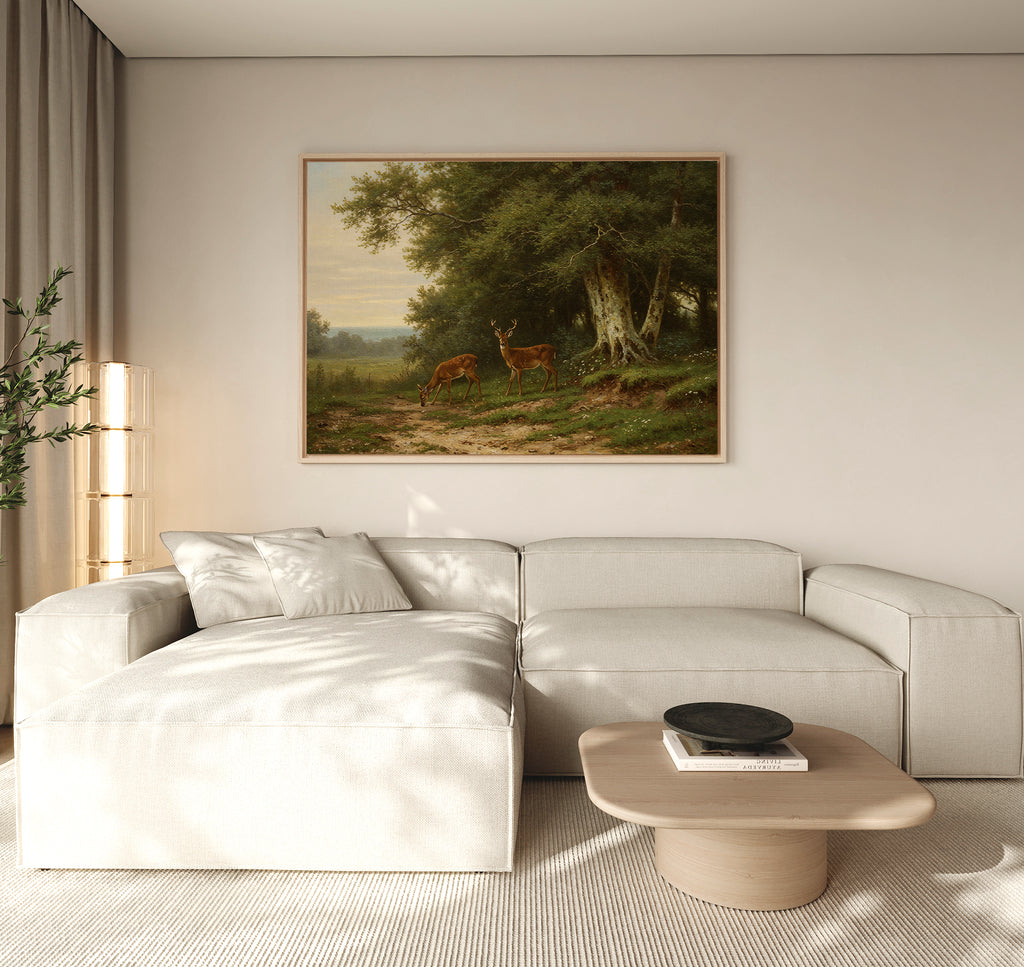 A minimalist living room features a light beige sectional, wooden coffee table, and the Woodland Watch Canvas Art depicting deer in a forest. Natural light filters through sheer curtains, with a green plant in the corner—ideal for nature lovers.