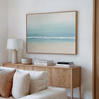 A minimalist living room features a light wood sideboard, white lamp, stacked books, and a neutral-toned bowl. Above hangs the Airy Shore Canvas Art - Horizontal, creating a calming coastal vibe. A beige sofa is placed in the foreground.