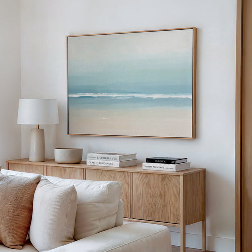 A minimalist living room features a light wood sideboard, white lamp, stacked books, and a neutral-toned bowl. Above hangs the Airy Shore Canvas Art - Horizontal, creating a calming coastal vibe. A beige sofa is placed in the foreground.