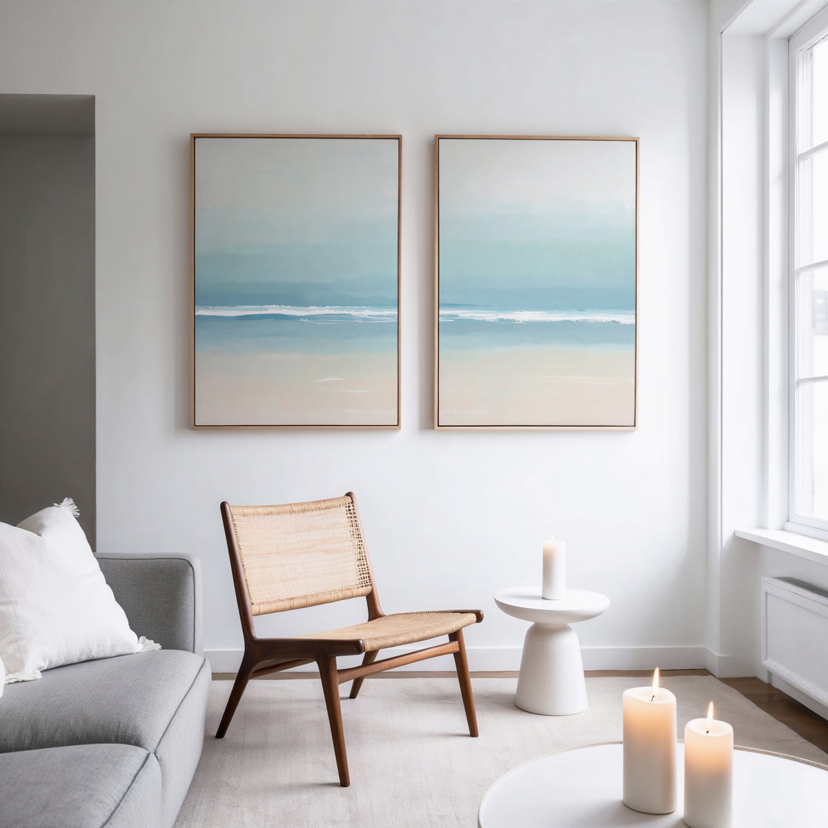 A bright, minimalist living room features a wooden chair, small white side table, and lit candles. Above the chair hangs the Airy Shore - Set Of 2 prints, bringing a calming coastal vibe as natural light fills the space.