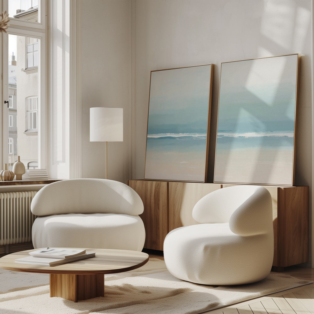 A bright, modern living room features two white rounded chairs, the Airy Shore - Set Of 2, paired with a low wooden coffee table and serene coastal print artwork on a wooden sideboard. Sunlight streams through large windows.