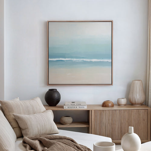 A minimalist living room with a beige sofa, neutral cushions, and a wooden sideboard. Above, the Airy Shore Canvas Art - Square creates a Nordic vibe, while decorative vases and books are neatly arranged on the sideboard.