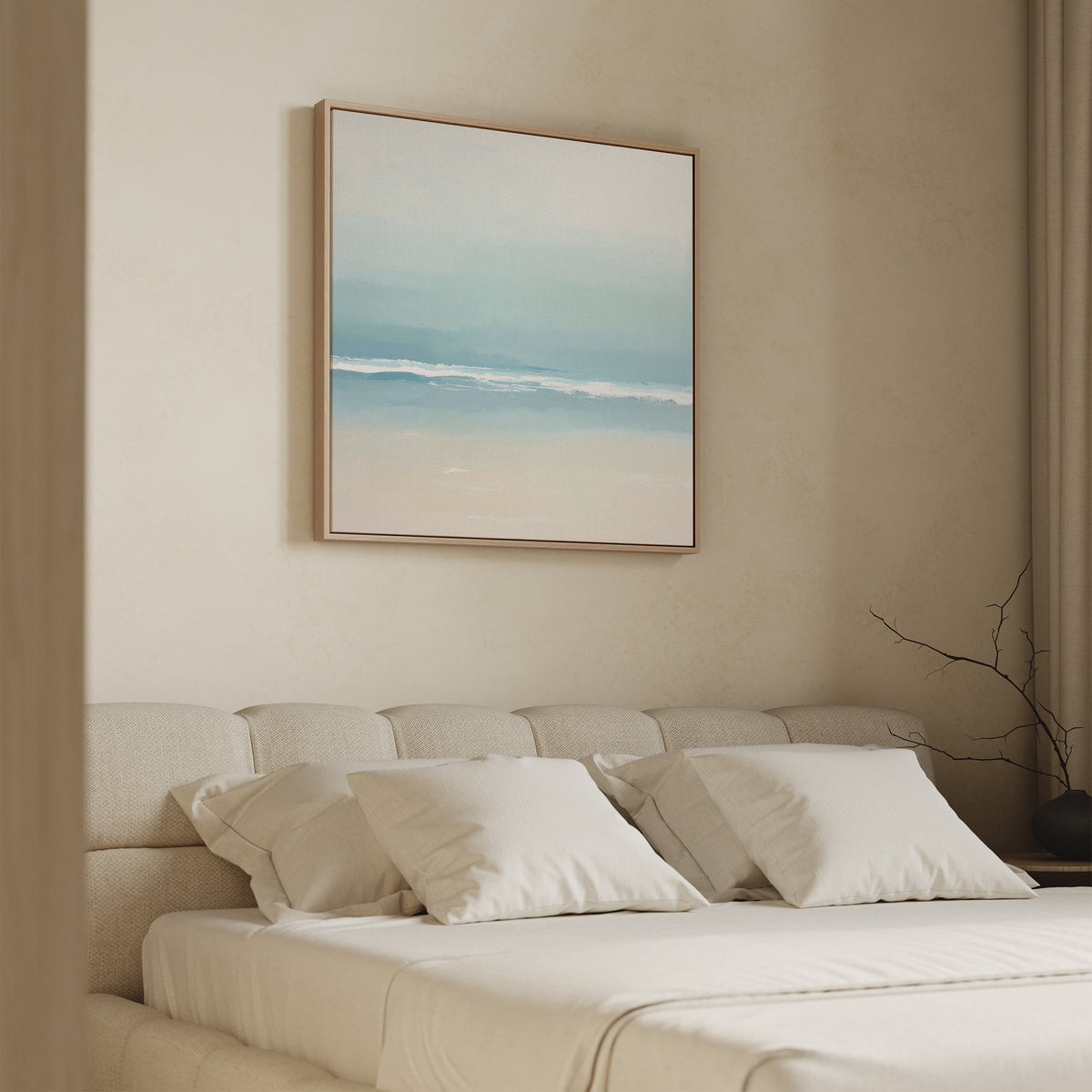 A minimalist bedroom features a beige upholstered bed with white bedding and pillows. Above the bed hangs the Airy Shore Canvas Art - Square, showcasing a soft abstract beach scene. A bare branch accents the bedside table.