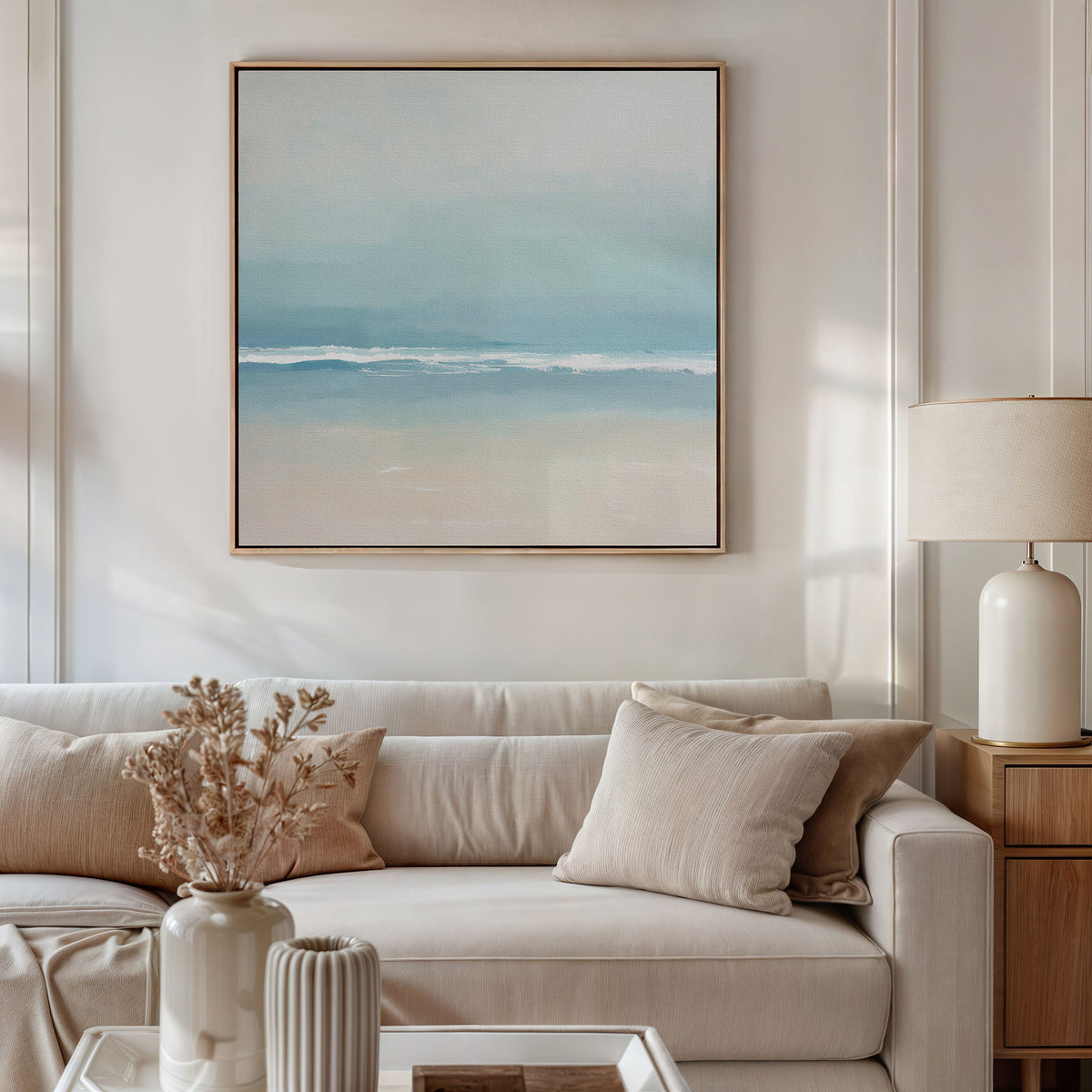 A beige sofa with cream pillows sits against a wall, above which hangs the Airy Shore Canvas Art - Square. A white table lamp and dried flowers in vases accent the tables, creating relaxing seaside decor.