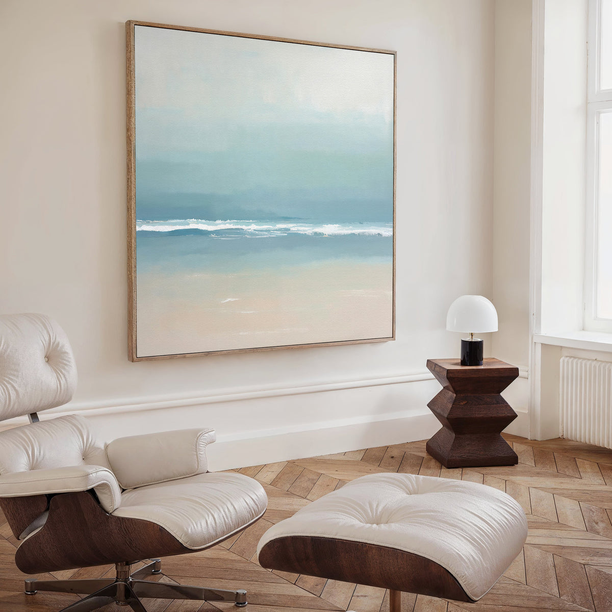 A minimalist room features a modern white lounge chair and ottoman, a dark wood side table with a white lamp, and the Airy Shore Canvas Art - Square, creating subtle seaside decor as sunlight streams through a tall window.