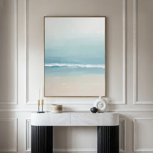 A minimalist room with a white paneled wall, Airy Shore Canvas Art - Vertical in an abstract seascape style, and a marble console table adorned with two candles, a round white sculpture, a small black bowl, and a wooden container.