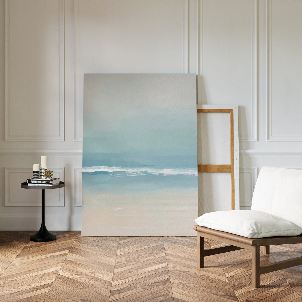 The Airy Shore Canvas Art - Vertical, featuring soft blue and beige tones of the sea, leans against a white paneled wall. A cushioned wooden chair and candles on a round table enhance the cozy, modern seaside vibe.