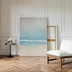 The Airy Shore Canvas Art - Vertical, featuring soft blue and beige tones of the sea, leans against a white paneled wall. A cushioned wooden chair and candles on a round table enhance the cozy, modern seaside vibe.