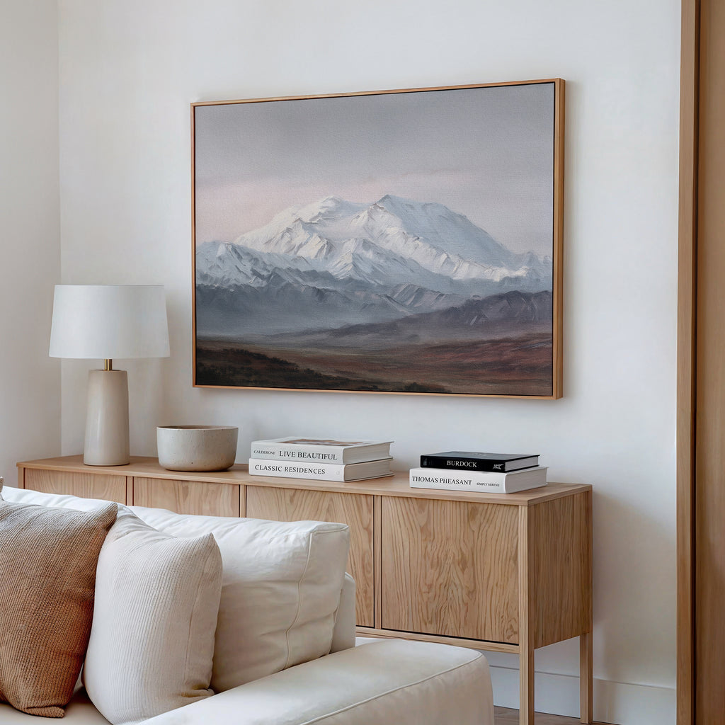 A minimalist living room features a wooden sideboard with stacked books, a white lamp, and a bowl. Above hangs the Alpine Majesty Canvas Art - Horizontal in mountain tones. A light-colored sofa in front adds subtle Nordic Park vibes.
