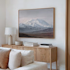 A minimalist living room features a wooden sideboard with stacked books, a white lamp, and a bowl. Above hangs the Alpine Majesty Canvas Art - Horizontal in mountain tones. A light-colored sofa in front adds subtle Nordic Park vibes.