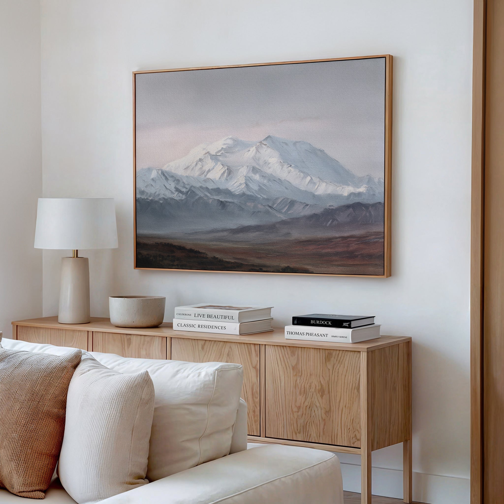 A minimalist living room features a wooden sideboard with stacked books, a white lamp, and a bowl. Above hangs the Alpine Majesty Canvas Art - Horizontal in mountain tones. A light-colored sofa in front adds subtle Nordic Park vibes.