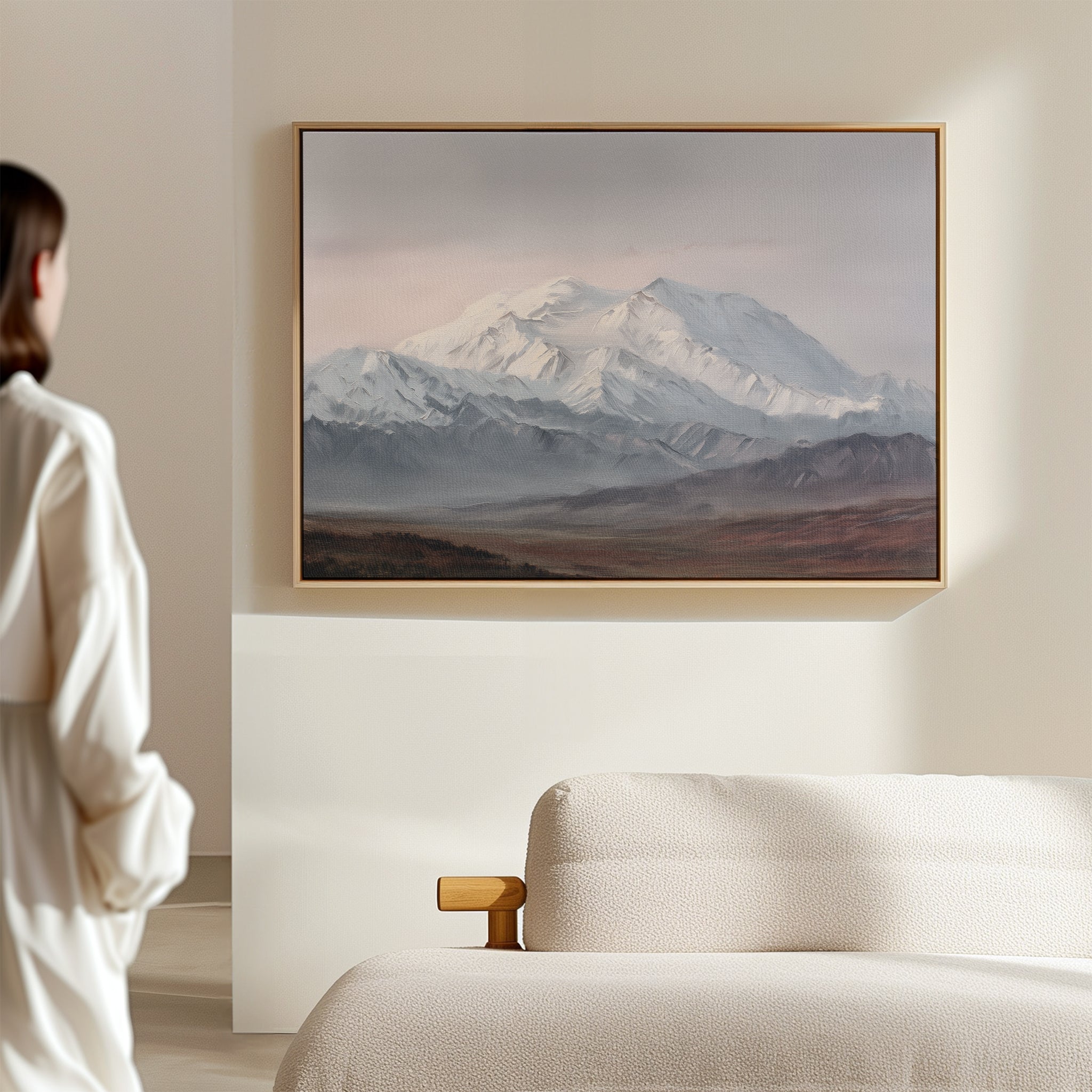 A person in white admires the Alpine Majesty Canvas Art - Horizontal, featuring snowy peaks and a Nordic Park palette, displayed above a light sofa in a minimalist room.