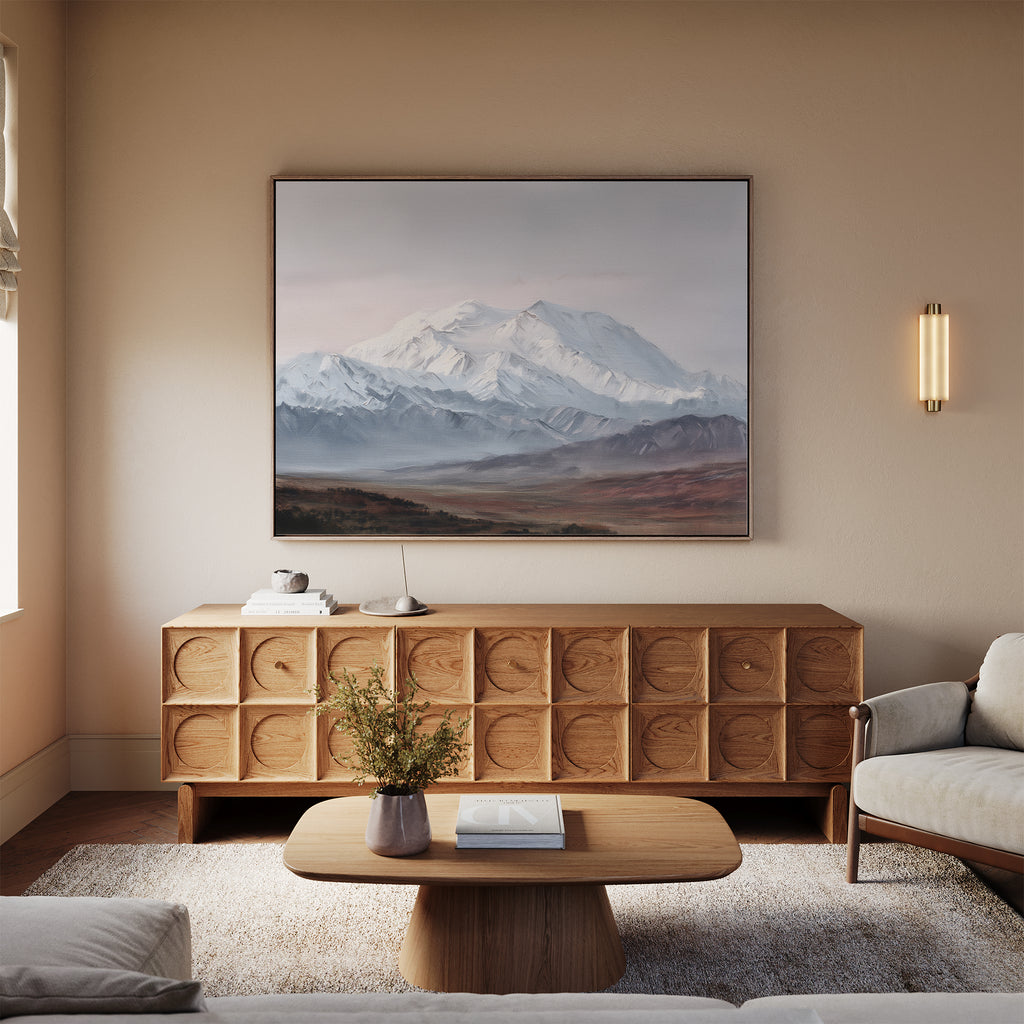 A cozy living room with a modern wood sideboard, low coffee table, and light-colored sofa features the Alpine Majesty Canvas Art - Horizontal, showcasing a balanced mountain palette. A small vase with flowers and books sit on the table.