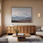 A cozy living room with a modern wood sideboard, low coffee table, and light-colored sofa features the Alpine Majesty Canvas Art - Horizontal, showcasing a balanced mountain palette. A small vase with flowers and books sit on the table.