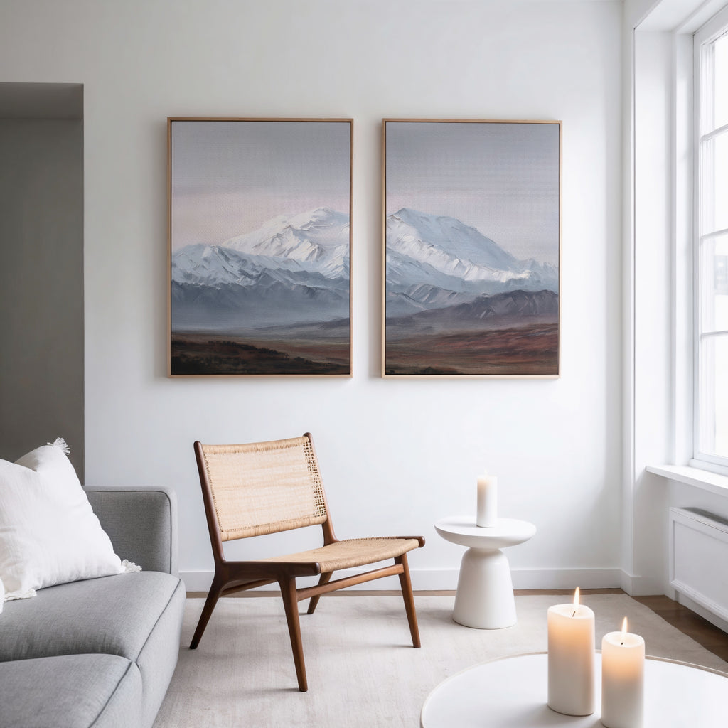 A modern, minimal living room features a woven chair, round white side table with a lit candle, gray sofa, and Alpine Majesty - Set of 2 mountain wall art. Three candles by the window complete the cozy Nordic Park atmosphere.