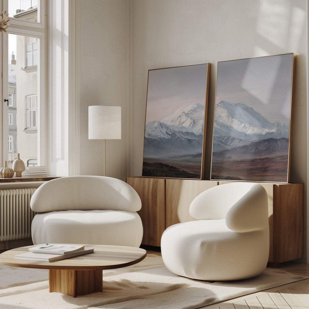A modern living room with two rounded white armchairs, a low wooden coffee table, a tall floor lamp, and the Alpine Majesty - Set of 2 mountain wall art leaning against a wooden sideboard by a sunny window for a serene Nordic feel.