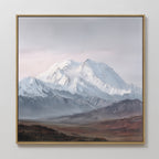 The Alpine Majesty Canvas Art - Square features a snow-capped peak beneath clouds, with rugged brown foothills and misty tones, adding a touch of mountain grandeur to any plain white wall.