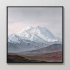 The Alpine Majesty Canvas Art - Square features a framed print of a snow-capped mountain under cloudy skies, with rocky foothills and a brown plain in front, displayed on a light wall.