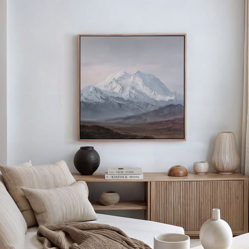 A minimalist living room with a beige couch, neutral cushions, wooden sideboard, decorative vases, and a large Alpine Majesty Canvas Art - Square displayed on a white wall.
