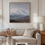 A cozy living room features a beige sofa with neutral pillows, a side table with a white lamp, and an Alpine Majesty Canvas Art - Square above the sofa. A coffee table holds a vase filled with dried flowers.