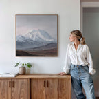 A woman in a white blouse and jeans stands by a wooden cabinet, looking at the Alpine Majesty Canvas Art - Square on the white wall. A plant and a closed book sit on top of the cabinet.