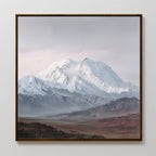 The Alpine Majesty Canvas Art - Square showcases a snow-capped mountain range under a cloudy sky, rugged foothills, and brown foreground terrain, beautifully displayed on a plain white wall.