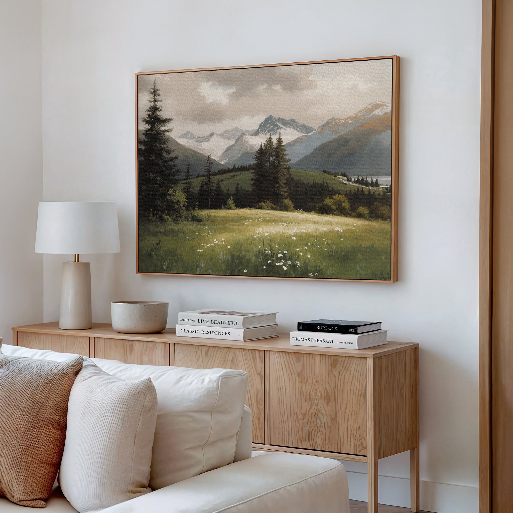 A modern living room features a light wood console, white lamp, and books, with the Alpine Meadow Canvas Art - Horizontal depicting a mountain landscape above the white sofa.