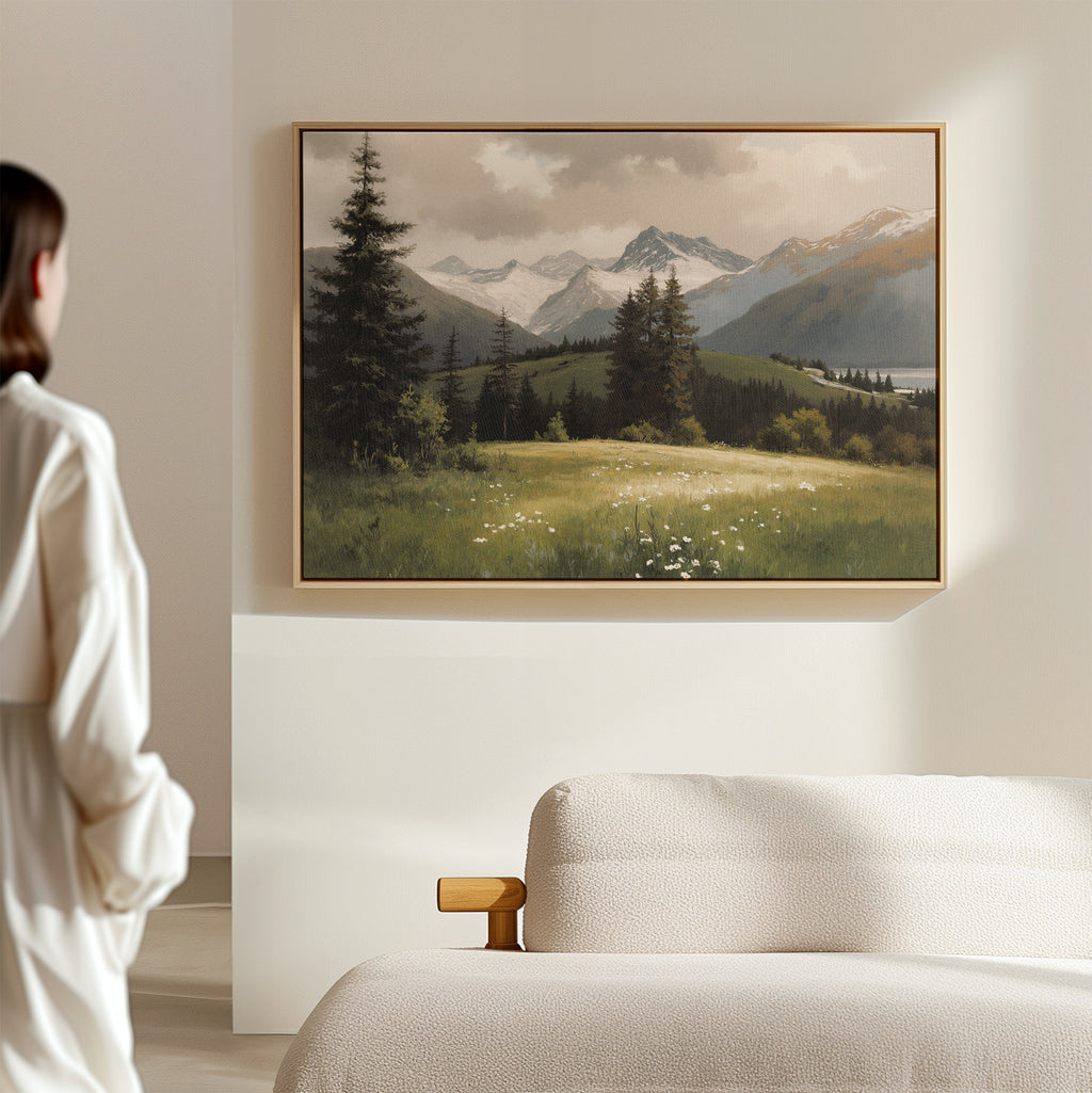 A person in a white shirt stands in a minimalist room, gazing at the Alpine Meadow Canvas Art - Horizontal featuring mountain scenery and pine trees, which hangs on a beige wall above a light-colored sofa.