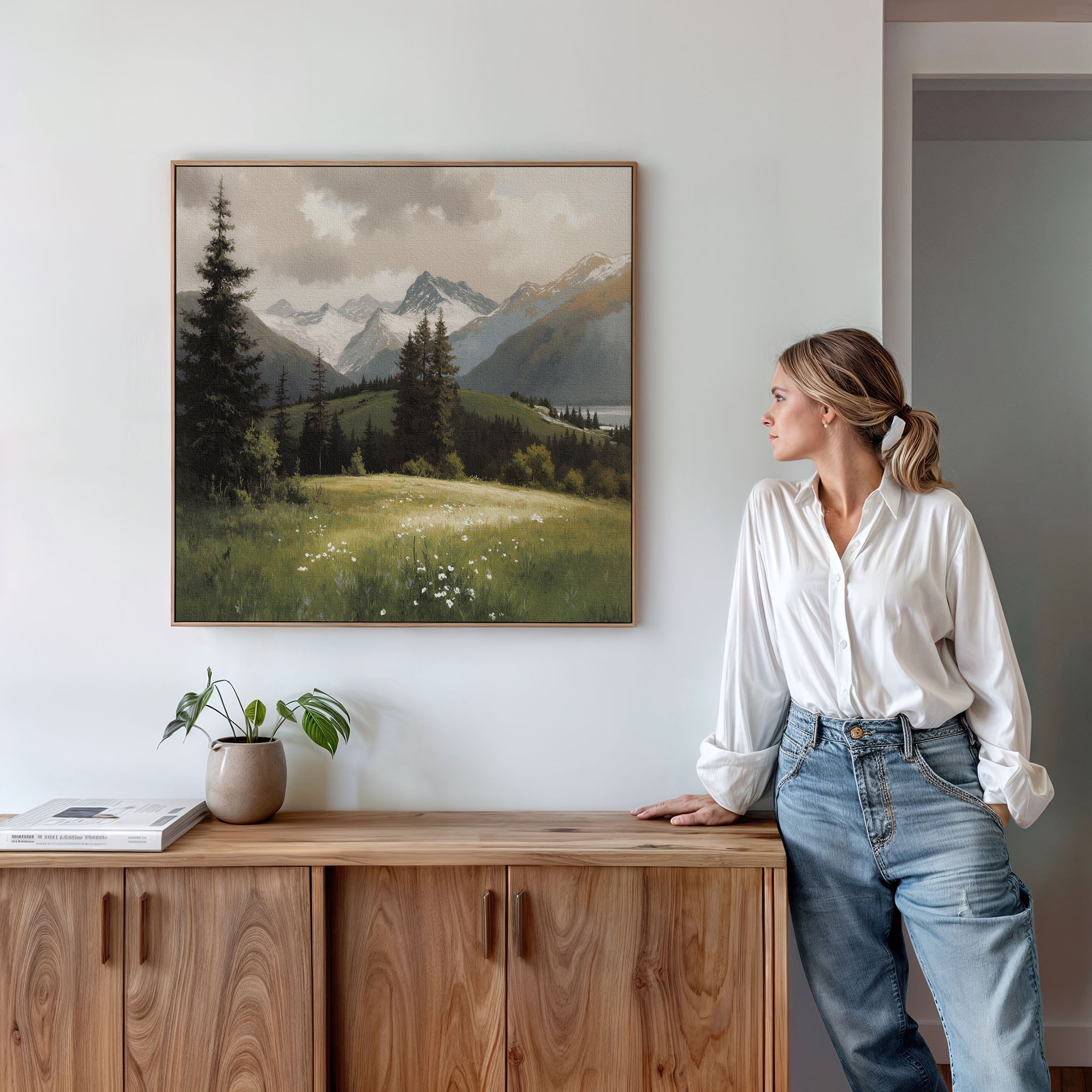 A woman in a white blouse and jeans leans on a wooden cabinet, gazing at the Alpine Meadow Canvas Art - Square hanging on the white wall above. A potted plant and a book sit on the cabinet.