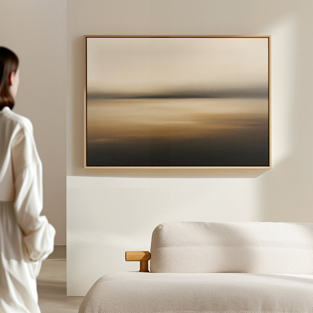 A person stands in a minimal, softly lit room with a textured beige sofa and the Amber Fields Canvas Art - Horizontal, a large abstract painting in warm amber tones, hanging on the wall.