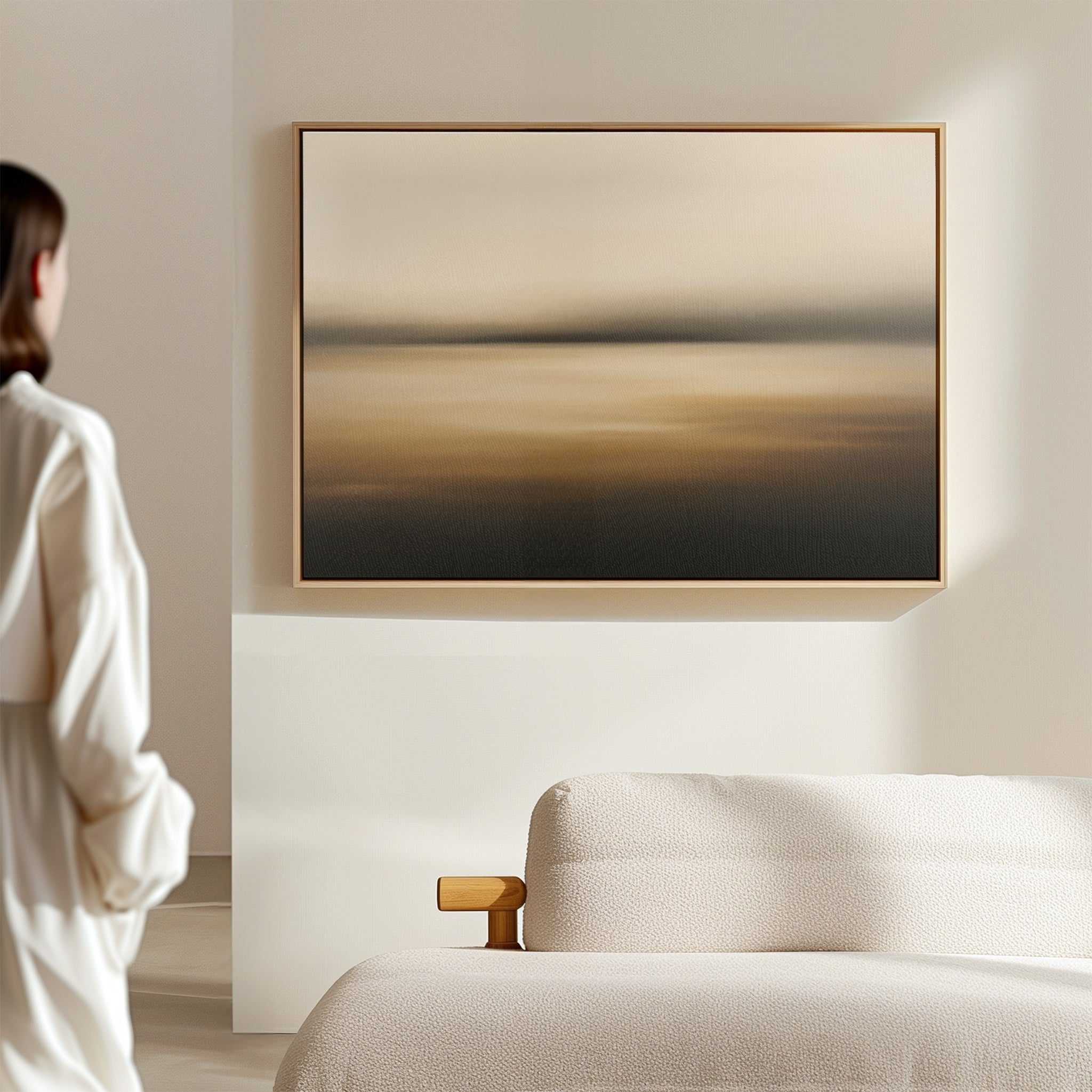 A person stands in a minimal, softly lit room with a textured beige sofa and the Amber Fields Canvas Art - Horizontal, a large abstract painting in warm amber tones, hanging on the wall.