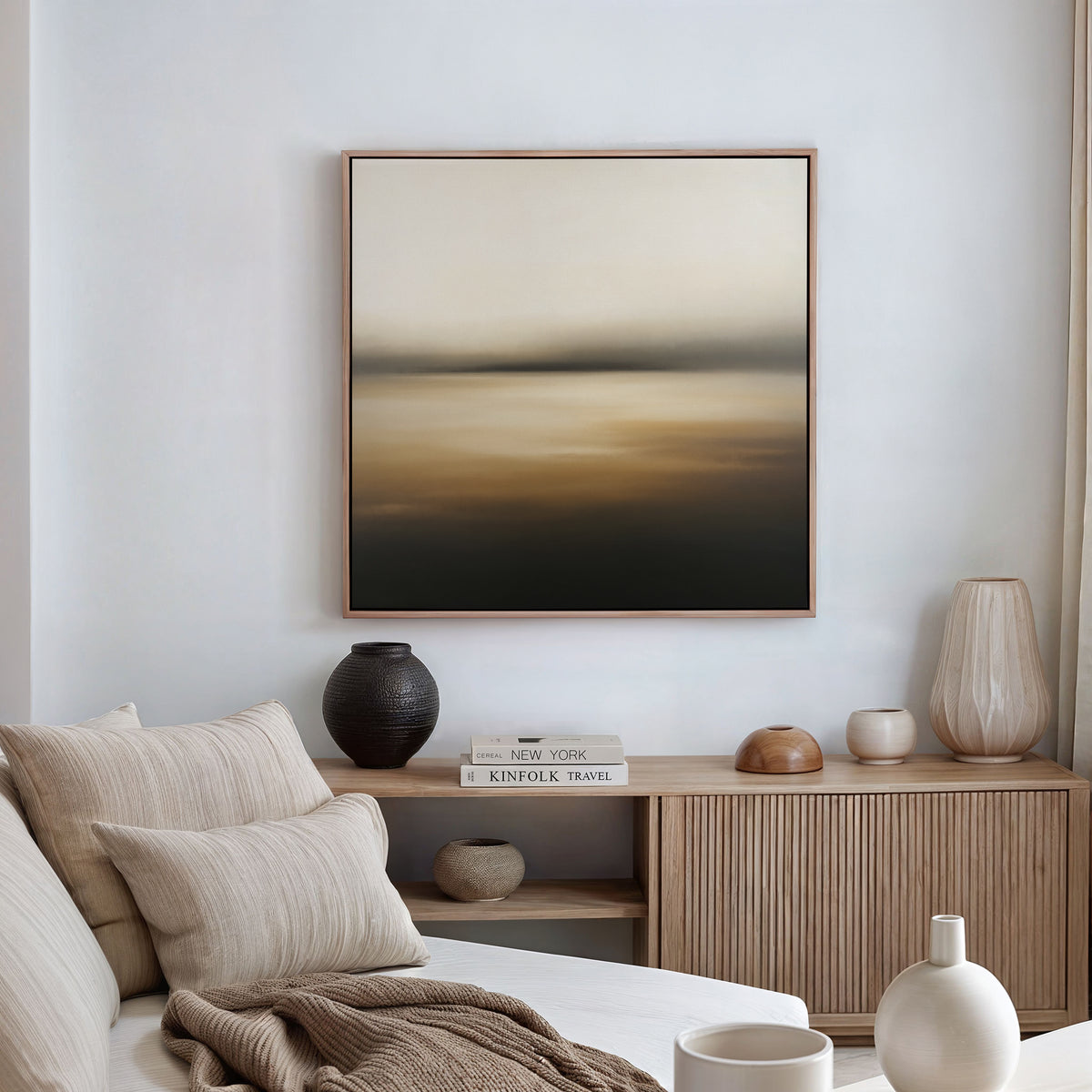 A serene living room features a beige sofa, neutral cushions, and a wooden sideboard with vases and books. Above, the Amber Fields Canvas Art - Square in soft amber and beige tones sets a tranquil mood.