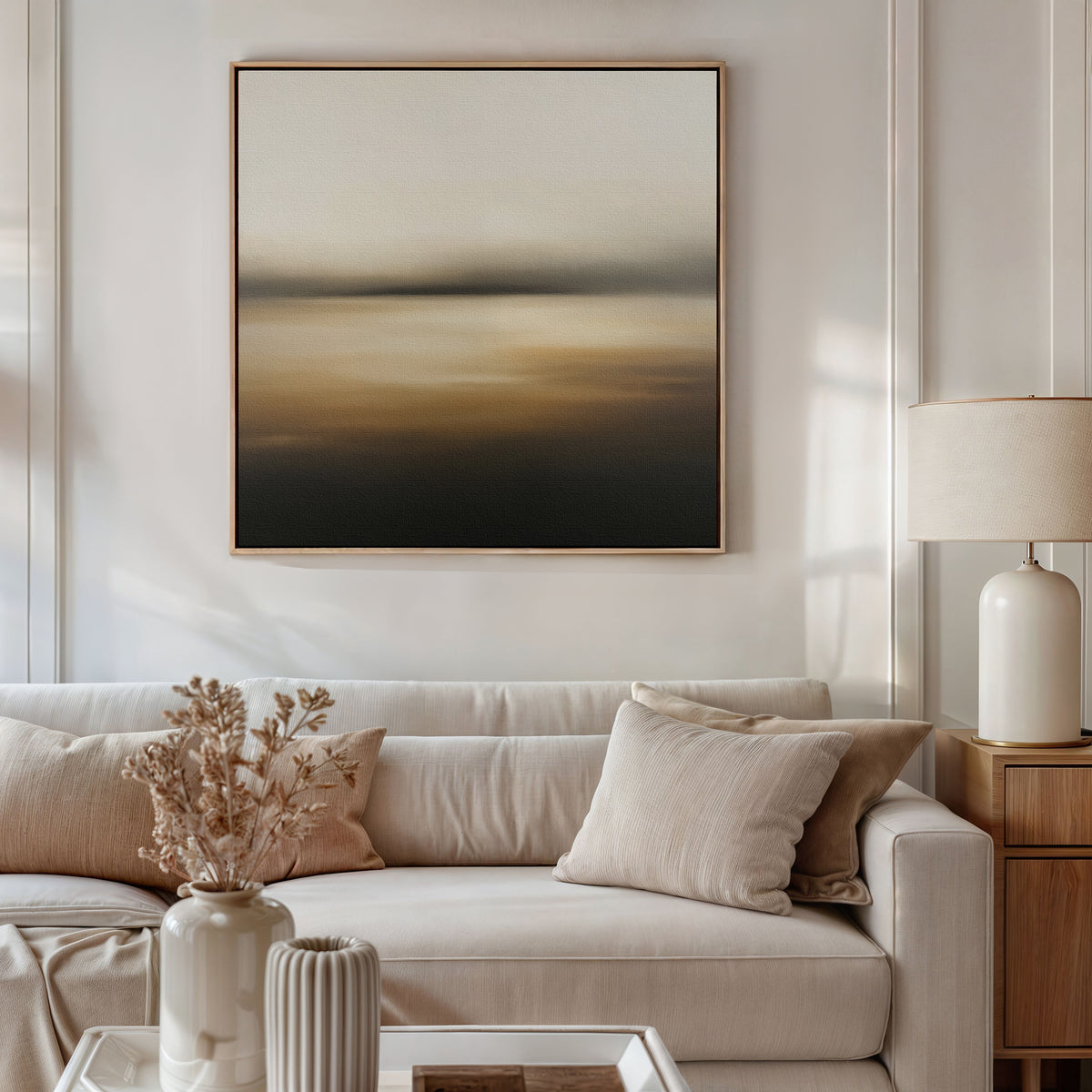 A neutral-toned living room featuring a light beige sofa, cream and beige cushions, the Amber Fields Canvas Art - Square on the wall, a table lamp, and a decorative vase with dried flowers on the coffee table.