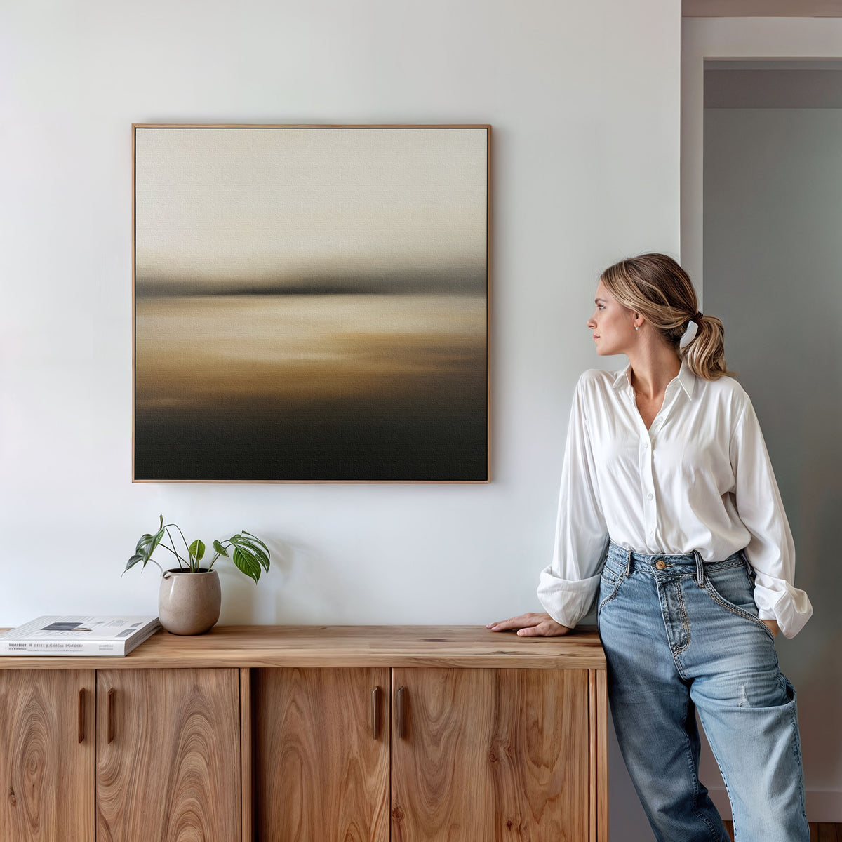 A woman in a white blouse and jeans stands by a wooden cabinet with a potted plant and books, admiring the Amber Fields Canvas Art - Square, an atmospheric landscape print with amber tones on a white wall.