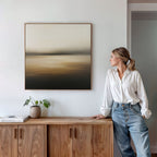 A woman in a white blouse and jeans stands by a wooden cabinet with a potted plant and books, admiring the Amber Fields Canvas Art - Square, an atmospheric landscape print with amber tones on a white wall.