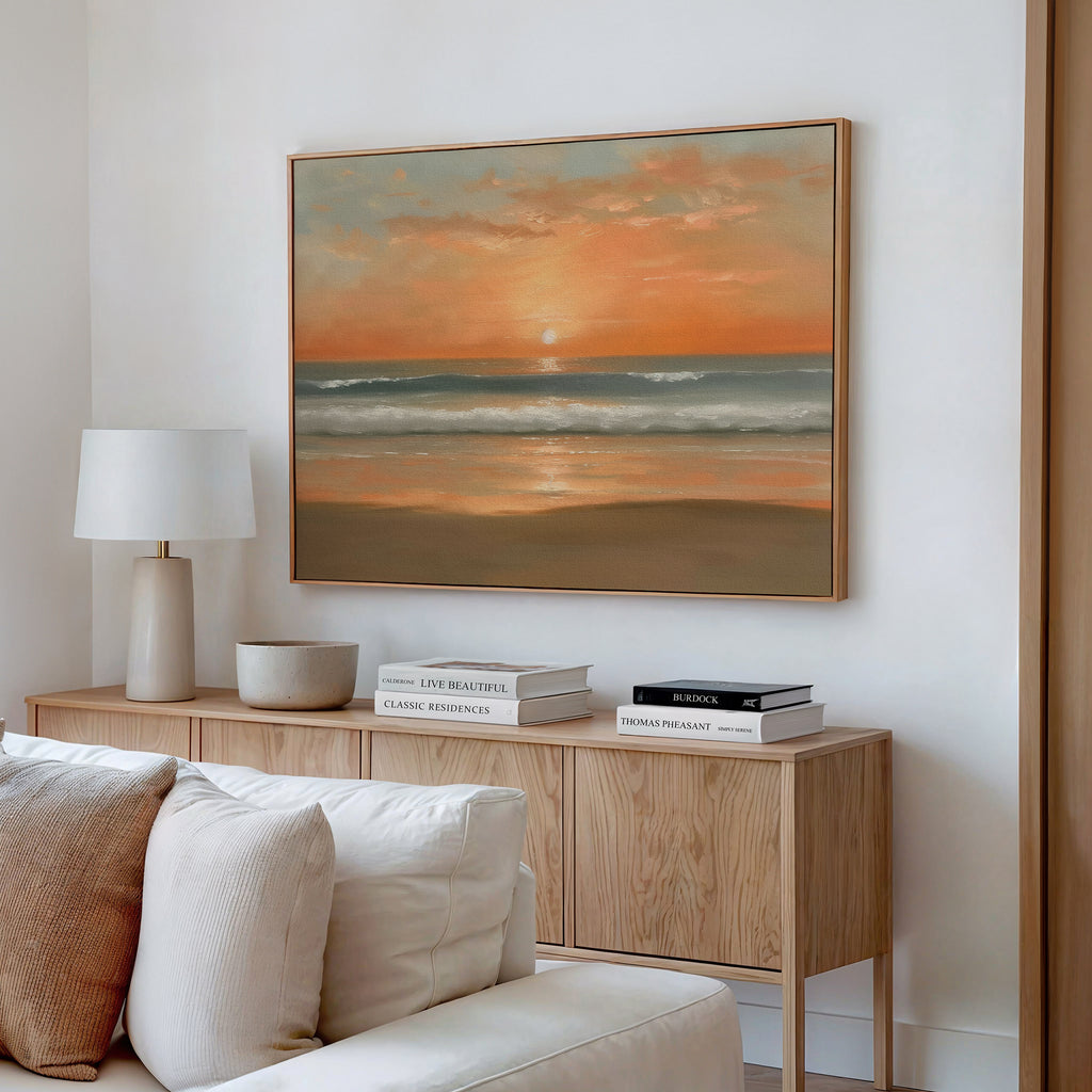 A modern living room with a wooden sideboard, stacked books, a table lamp, and the Amber Horizon Canvas Art - Horizontal depicting a coastal sunset over a calm ocean hanging on the white wall above.