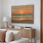 A modern living room with a wooden sideboard, stacked books, a table lamp, and the Amber Horizon Canvas Art - Horizontal depicting a coastal sunset over a calm ocean hanging on the white wall above.