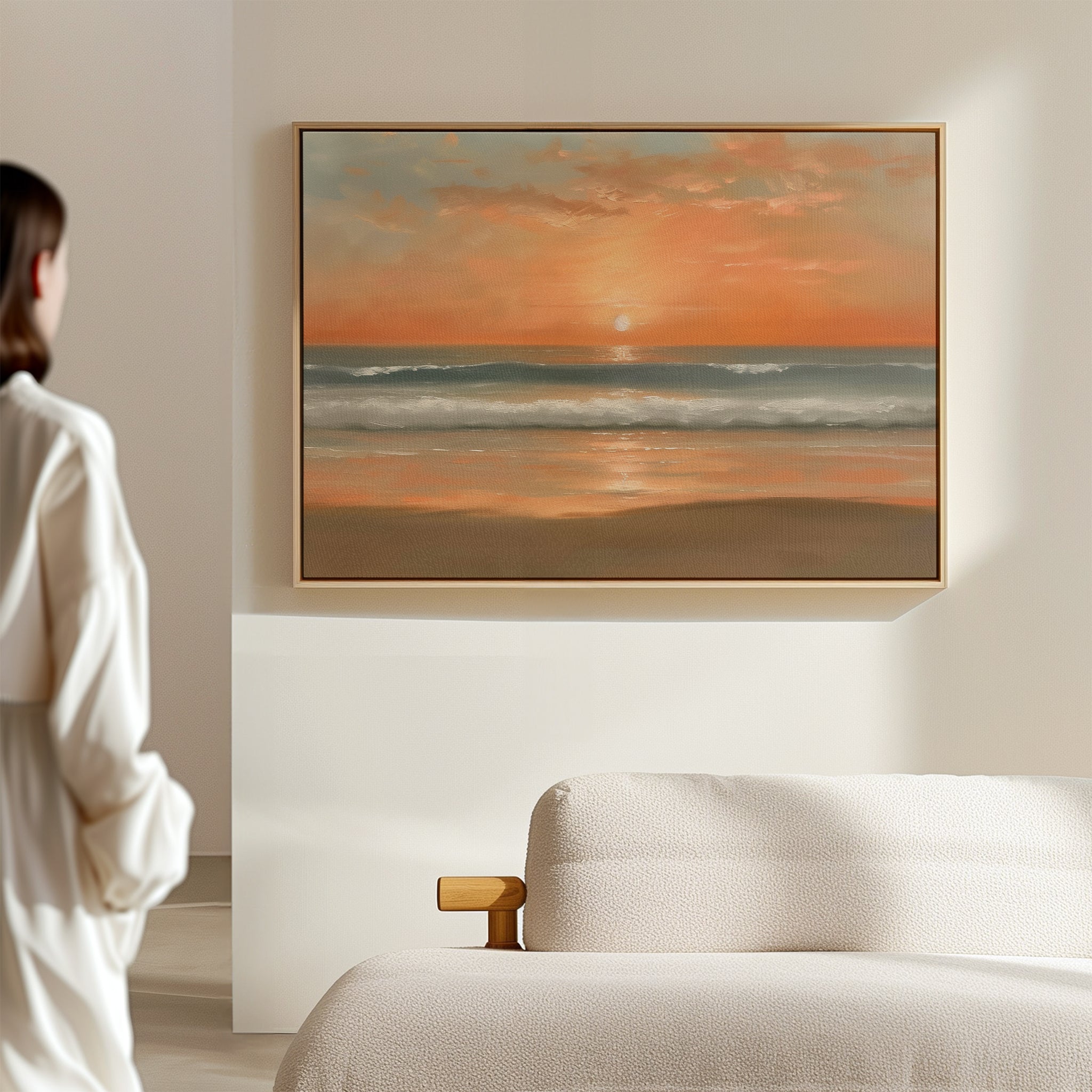 In a minimal, neutral-toned room, someone admires the Amber Horizon Canvas Art - Horizontal, which features an orange and pink coastal sunset over calm ocean waves with soft reflections.