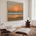 A modern living room with a white lounge chair and ottoman, wooden side table with a small lamp, and the Amber Horizon Canvas Art - Square featuring a coastal sunset with waves and an orange sky on the wall.