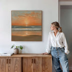 A woman in a white blouse and jeans stands by a wooden cabinet, admiring the Amber Horizon Canvas Art - Square on the white wall. A potted plant and books add charm to this Nordic Park-inspired setting.