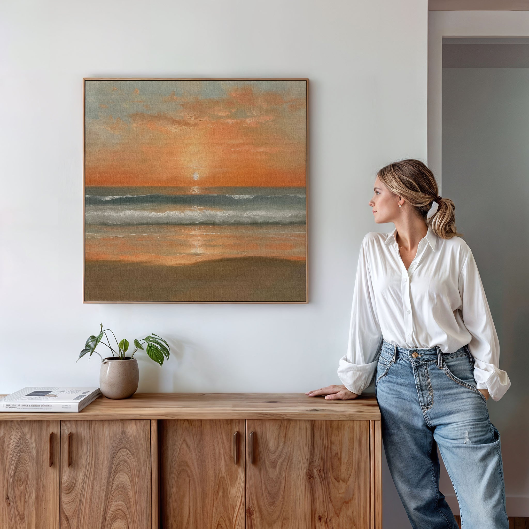 A woman in a white blouse and jeans stands by a wooden cabinet, admiring the Amber Horizon Canvas Art - Square on the white wall. A potted plant and books add charm to this Nordic Park-inspired setting.