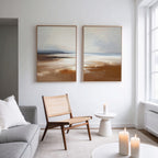 A modern, minimal living room features a light gray sofa, wooden chair, and the "Amber Shore - Set Of 2" abstract landscape paintings. A small white side table and lit candles add coziness as natural light streams through large windows.