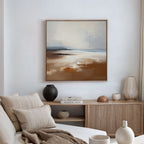 A modern living room in neutral tones features a beige sofa with cushions, a wooden sideboard with vases and books, and the Amber Shore Canvas Art - Square—an abstract landscape—displayed above as tranquil wall decor.