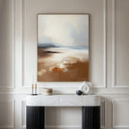 A modern console table with decorative accents stands against a paneled wall, topped by the Amber Shore Canvas Art - Vertical, featuring a tranquil abstract landscape in soft, earthy tones reminiscent of a serene beach scene.