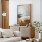 A modern living room featuring the Amber Shore Canvas Art - Vertical, a light wood sideboard, stacked books, ceramic vases, a small potted plant, a round floor lamp, and a white sofa with neutral cushions.