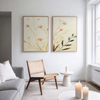 A modern, minimal living room features a light gray sofa, wicker chair, round white side table, and three lit candles. Above the chair hangs the "Autumn Botanicals - Set of 2" canvas prints in elegant beige tones. Natural light pours in from a large window.