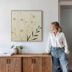 A woman in a white blouse and jeans stands by a wooden cabinet, admiring the Autumn Botanicals Canvas Art - Square with yellow flowers above her. A small plant and books sit on the cabinet.